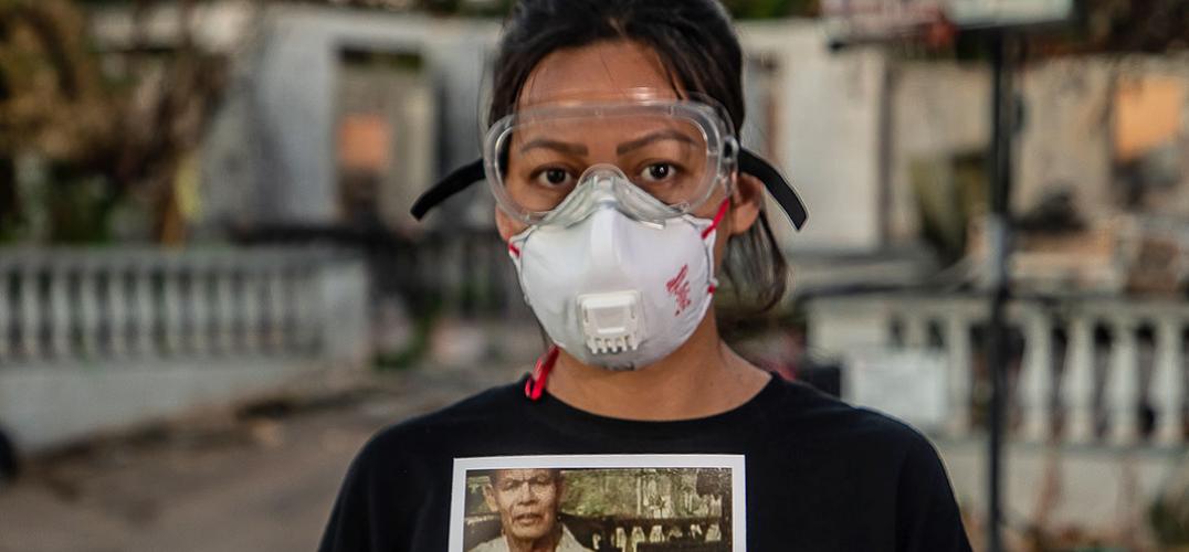 A woman wearing safety goggles and a respirator mask stands in front of the remains of her home holding a photograph