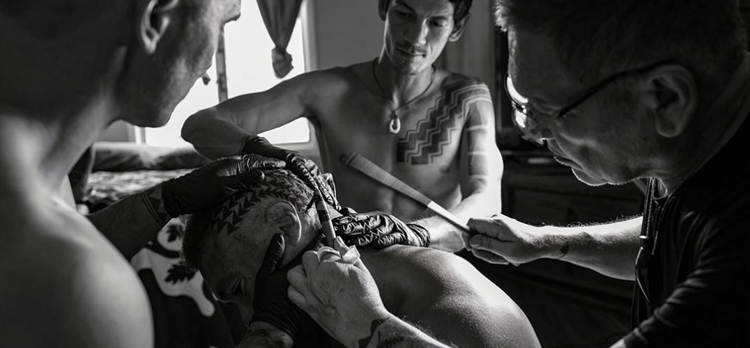 A tattoo artist applies a tattoo while two assistants stretch the skin of the recipient