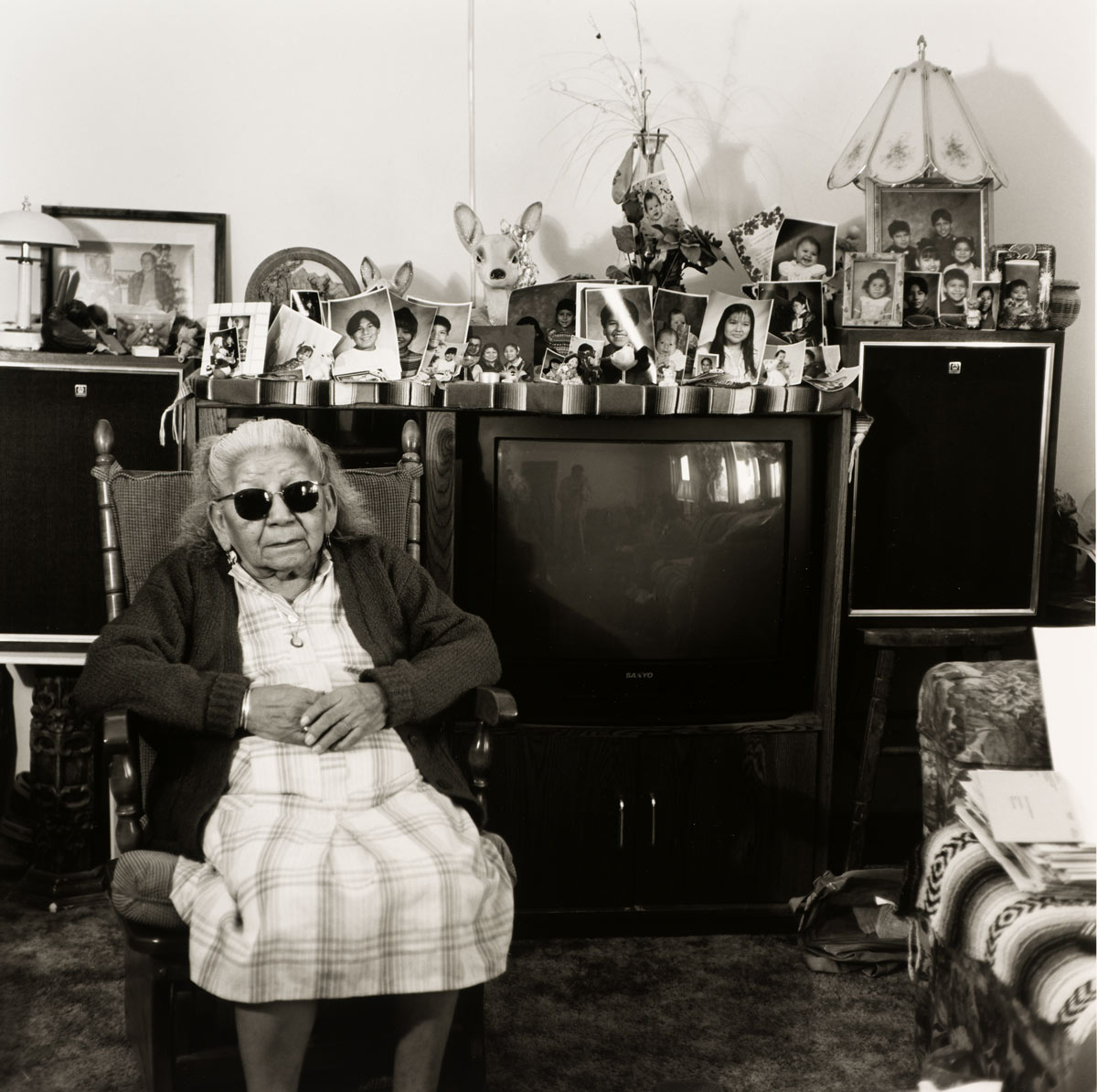 A woman in dark sunglasses sits in a rocking chair surrounded by family photographs.