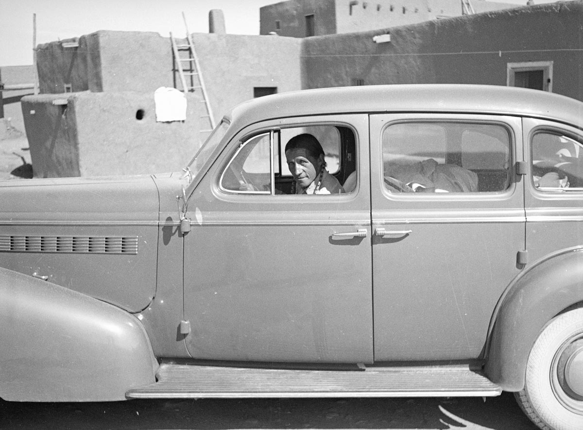 A man sits in the driver seat of a 1930s-era automobile.