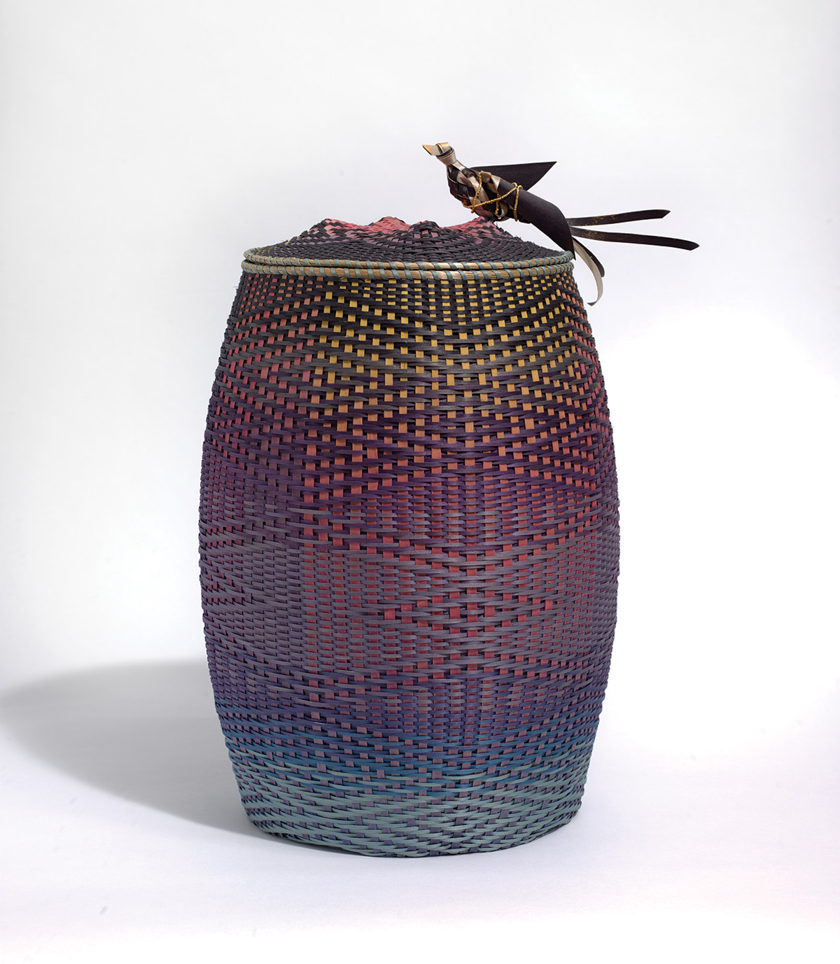 A tall, multicolor woven basket with a bird figure perched on its lid