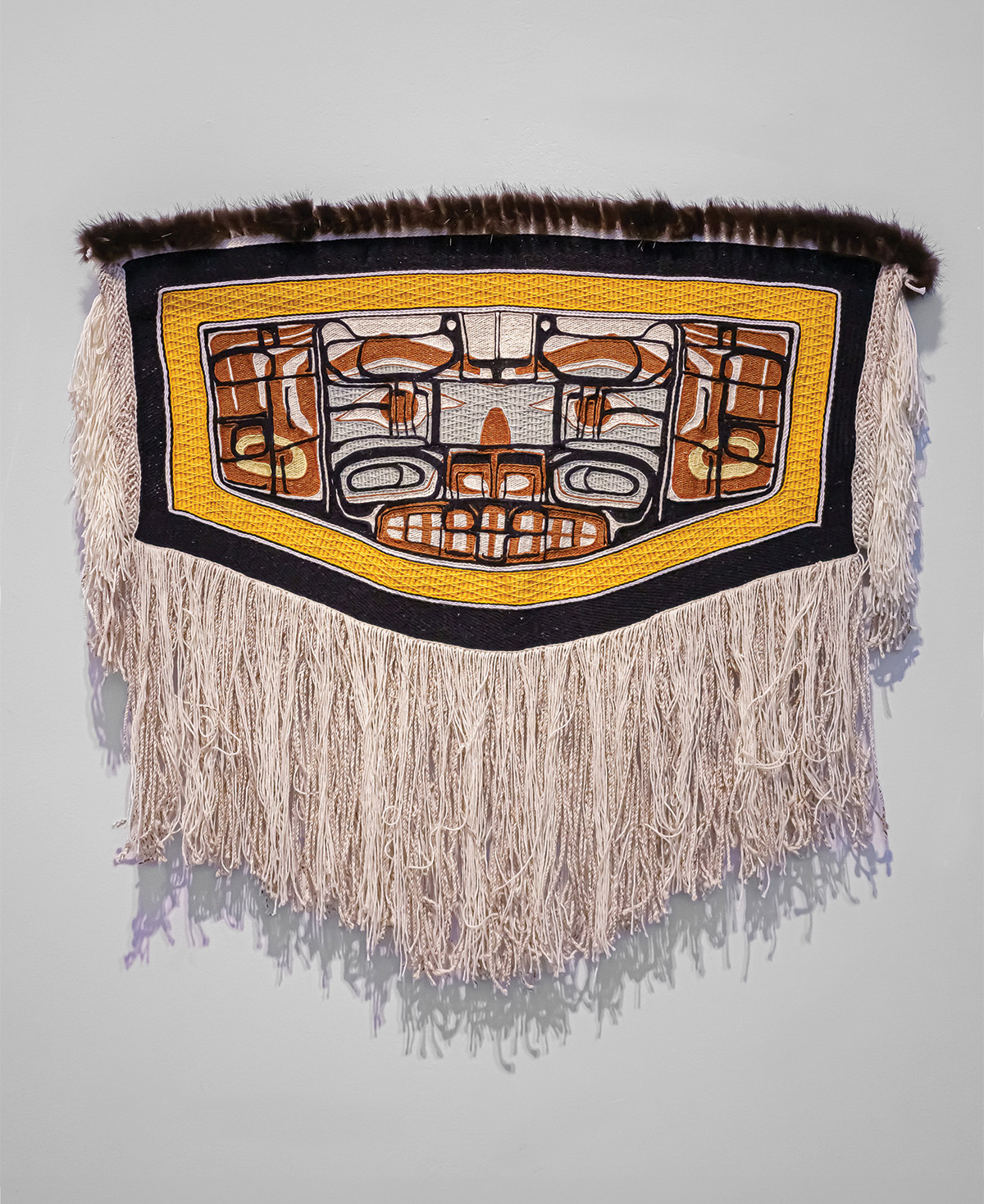 A woven robe with yellow, black, orange, grey, and white designs, brown beaver fur and a white fringe