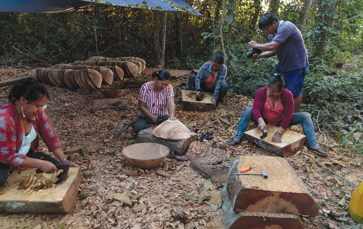 A group of people carving wood outdoors while being filmed by a filmmaker
