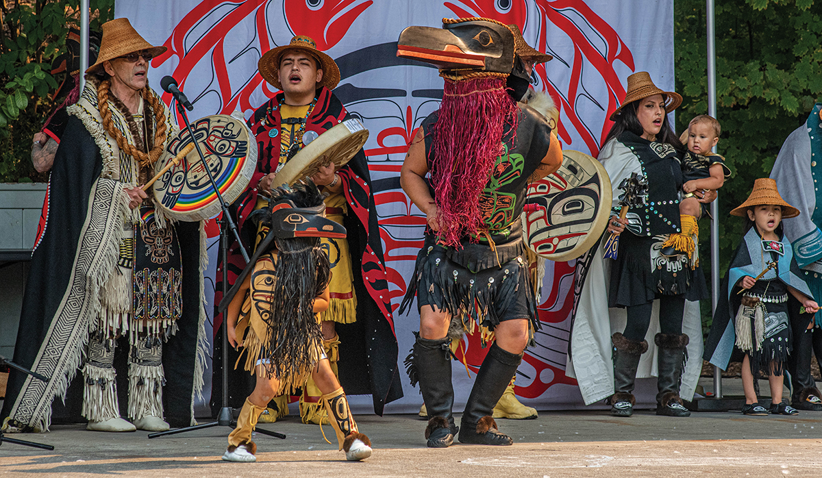 A group of performers on a stage sing, drum, and dance in regalia and costumes