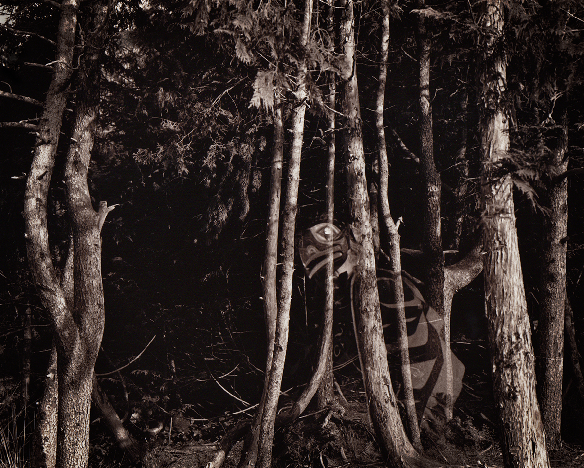 A black and white image of Tlingit carver wearing a Raven mask and regalia looks out from behind a tree in a forest