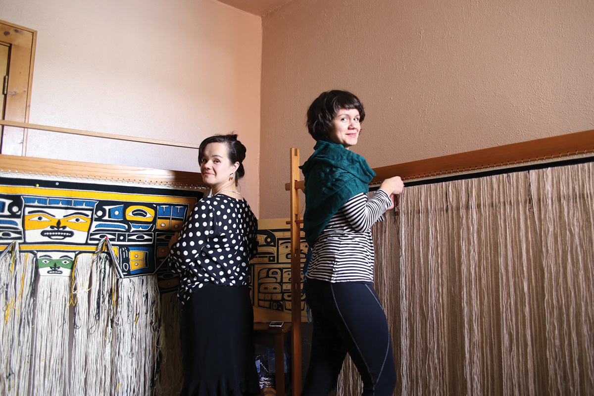 Lily Hope and Ursala Hudson stand back to back, each in front of a weaving loom