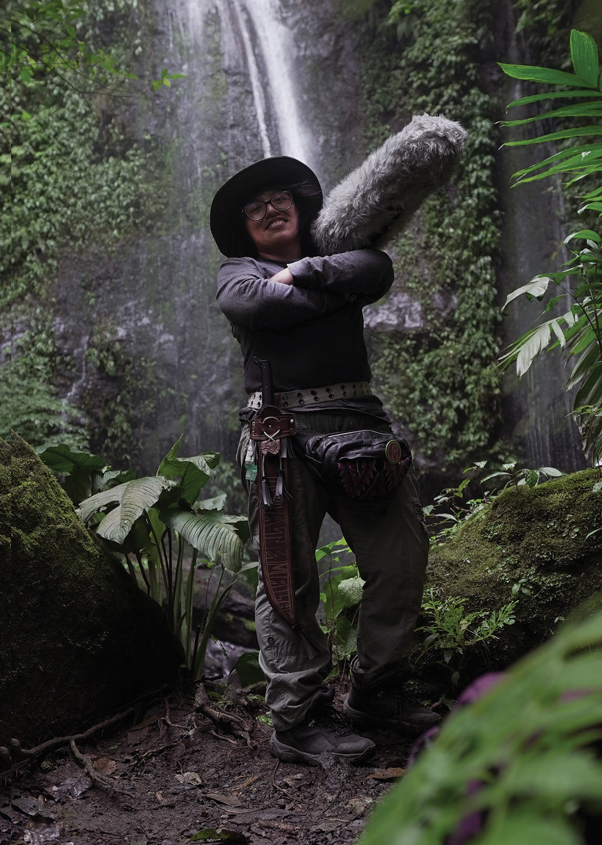 A person stands in front of a waterfall, carrying filmmaking equipment