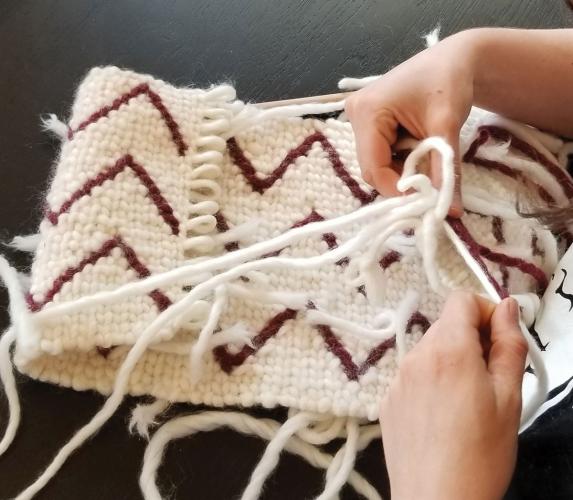 Two hands in the process of weaving a small white bag with red zigzag lines