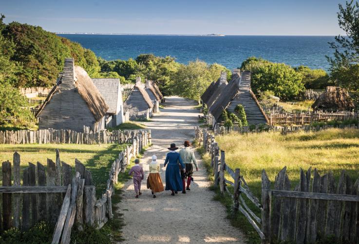 Four people in historical costume walk through the reconstructed village of New Plimoth