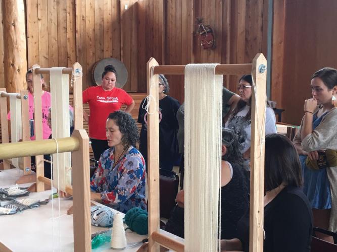 A person sits at a table and works with weaving equipment, while other people stand behind her and observe