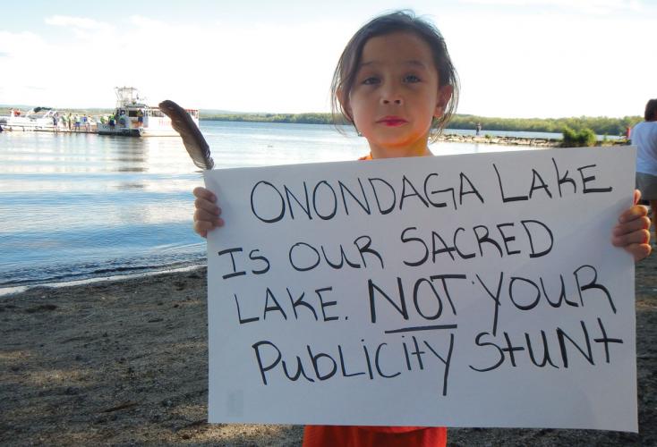 A child holds a protest sign that says "Onondaga is our sacred lake. Not your publicity stunt."