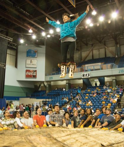 Dozens of people holding an animal-skin blanket toss a woman up into the air