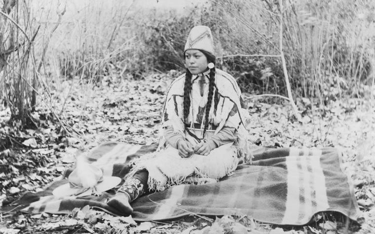 Cayuse tribal member Agnes Davis sits on a blanket amid plants. She is a wearing traditional Cayuse dress, jewelry and a fezlike hat woven from dogbane fibers.