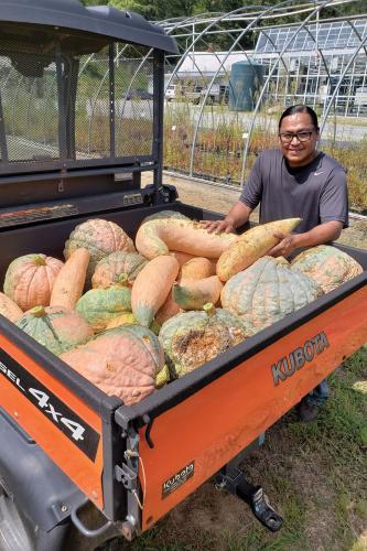 In North Carolina, a man stands next to a truck, the bed of which is filled with squash.  Behind him is a greenhouse and farm.