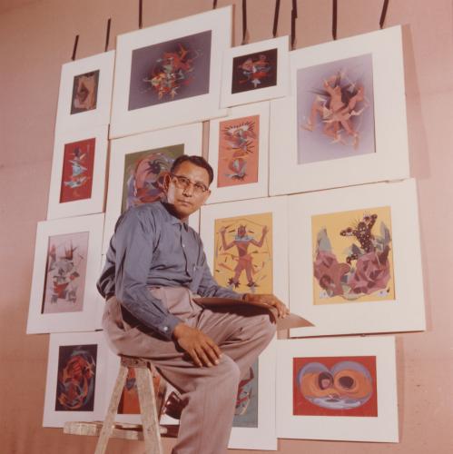 Portrait of Oscar Howe seated in front of a selection of his paintings hanging on a wall.