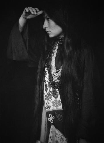 Zitkala-Ša, a Yankton Sioux woman wearing traditional clothing and jewelry, holds her hand to her forehead