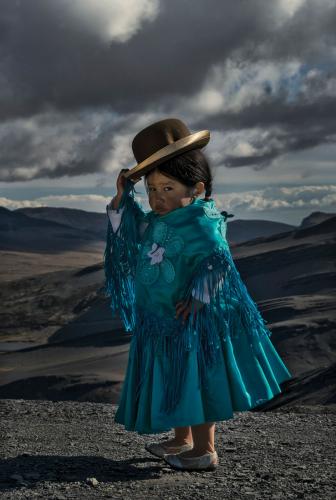 Nathaly Patzy, an Aymara girl in Bolivia wearing traditional cholita attire, tips her bowler hat as she looks at the photographer.