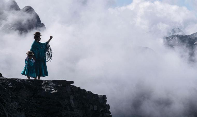 As her daughter holds her hand, an Aymara woman in traditional cholita attire reaches out over a foggy crevasse to the Andean mountains