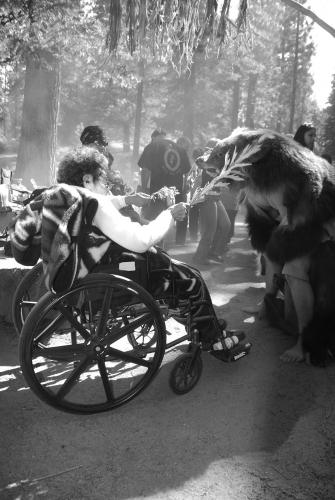 A black and white image. A woman in a wheelchair touches the bear skin with a sprig of wormwood.