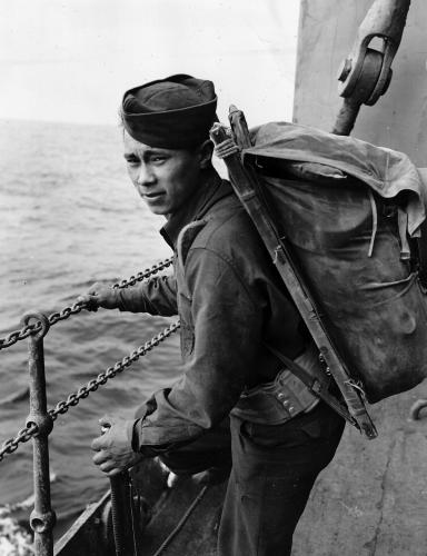 A young Aleut man in army uniform and gear looks at the camera while holding onto the chain rail of the military ship.