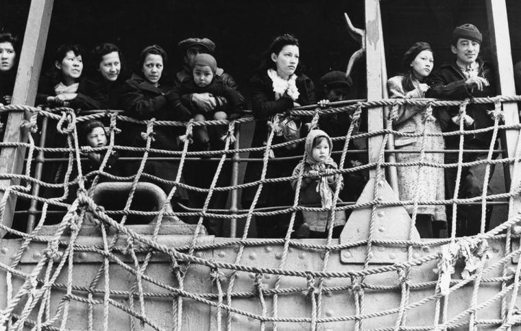 A group of Aleut residents of Pribilof Islands hold onto a railing and look off the ship. The image is in black and white.