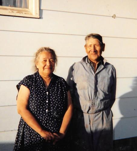 An Indigenous woman in a dark polka dot dress stands left of an Indigenous man in a matching gray jacket and pants. They are posed in front of a white siding with golden afternoon light.