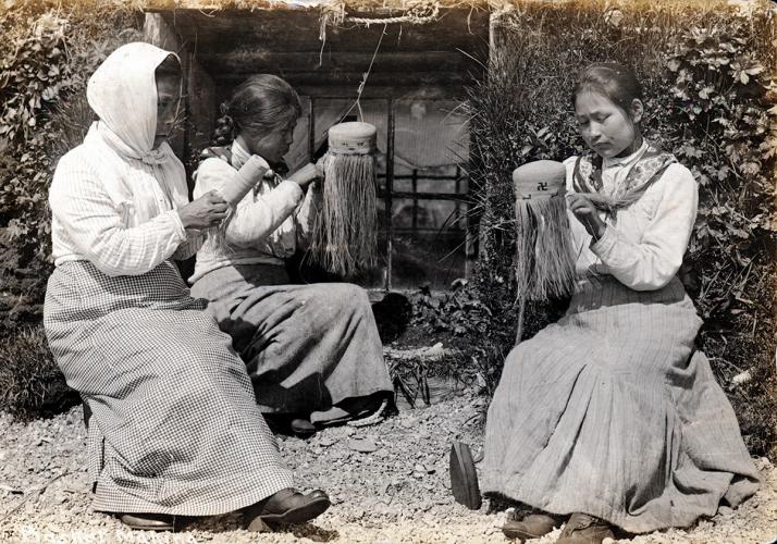 Three Aleut women sit outside of the entrance to a house that has been dug out of the hillside. They are each weaving a basket. The image is black and white.