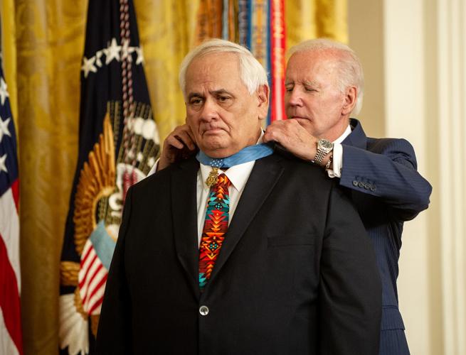 Dwight Birdwell wears a dark suit, white button-down, and red tie. President Biden, also wearing a suit, stands behind his back and places the blue-ribboned medal around Birdwell's neck.