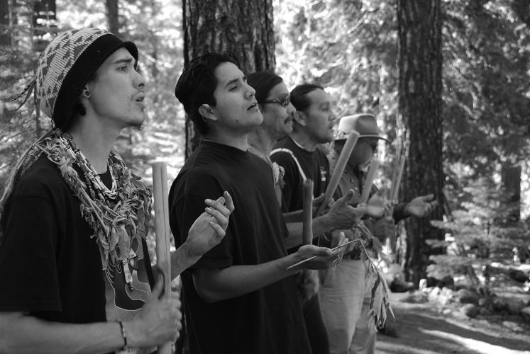 A black and white image of five men standing in a line in the forest, each singing and holding instruments.