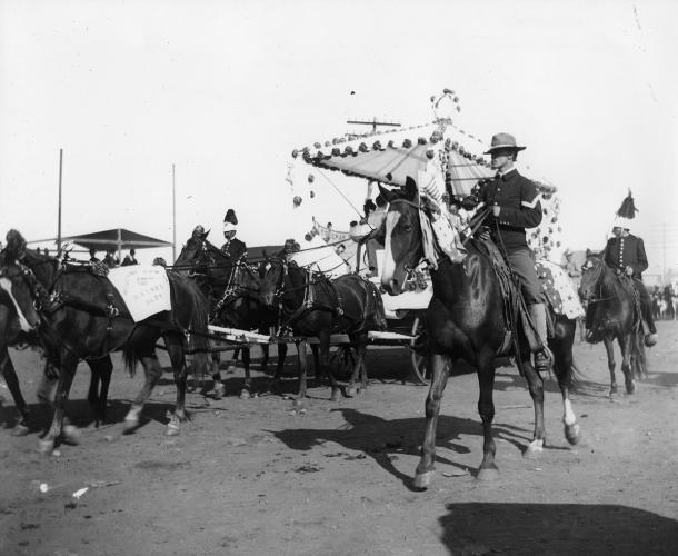 A black and white photograph of a parade procession with men in uniform on horseback pulling a decorated carriage.