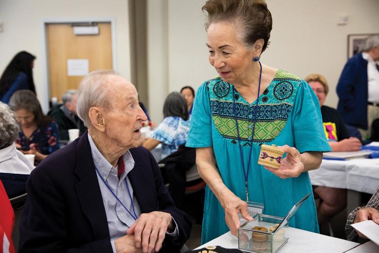An Attu Aleut descendant stands to the right of a U.S. veteran, holding a small woven basket and glass case with two more smaller baskets.