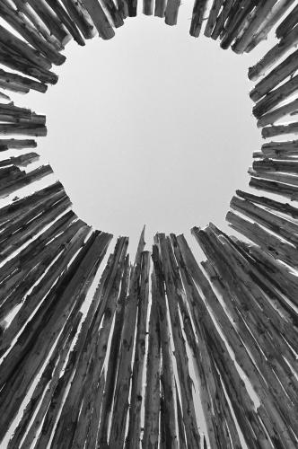 A black and white photo taken from inside a roundhouse, looking up towards the hole at the very top of the ceiling. The wooden beams create a circular opening to the sky.