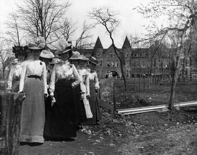 A group of well-dressed women in large frilly hats poses outside of a school building. The image is black and white.