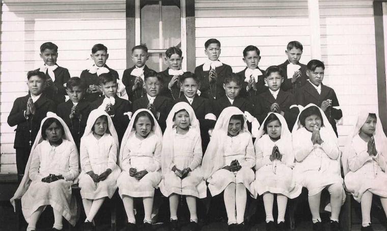 A black and white group photograph of Indigenous school children, some with their hands clasped in prayer. Girls dressed in all white sit in the front row, while boys dressed in all black stand behind them.