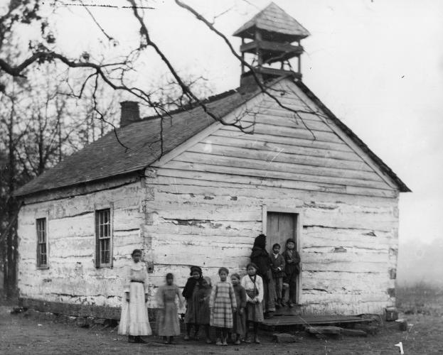 A black and white image of a rustic wooden single-room school house. A group of children and a young woman stand out front.