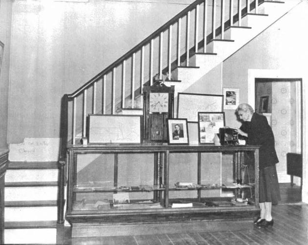 Jennie Cobb, now in her old age, stands to the right of a display case at the base of the stairs in Hunter Home. The photo is black and white.