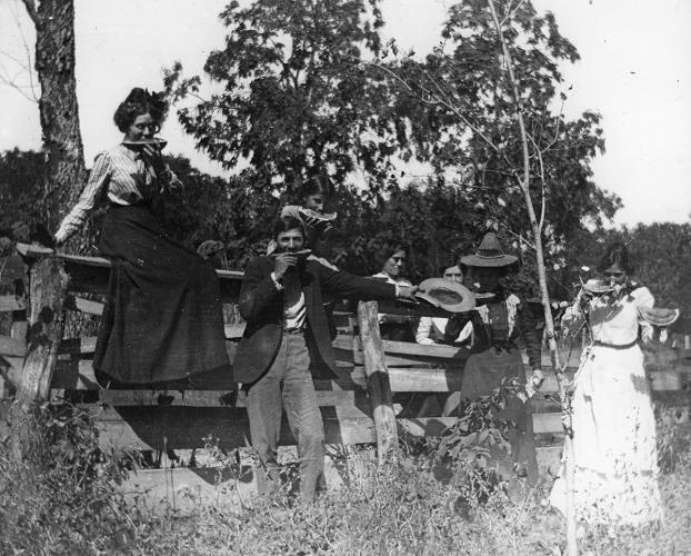A group of women and a man lean and sit against a wooden fence enjoying watermelon. The image is black and white.