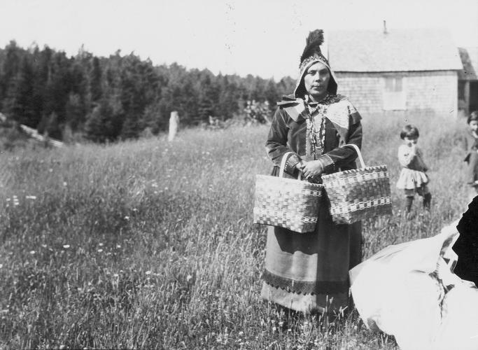 A black and white archival image of a woman with two baskets hanging from her arms and her young daughter in background