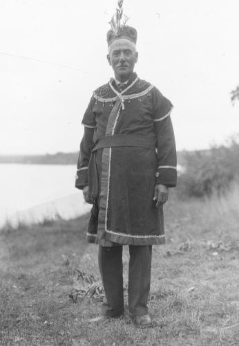 A black and white archival image of a man standing on a shore wearing a beaded coat and feathered band for a hat