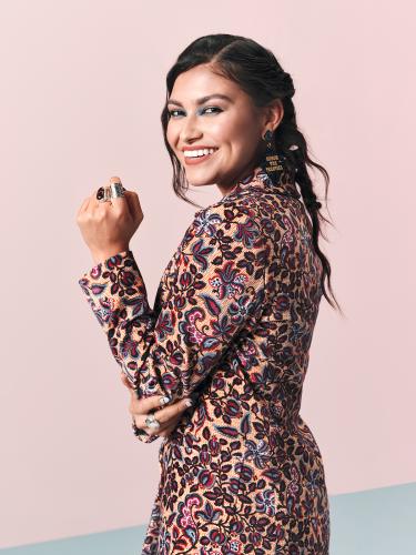 Actress Alyssa Wapanatâhk poses wearing rings created by Erik Lee in front of a pink backdrop.