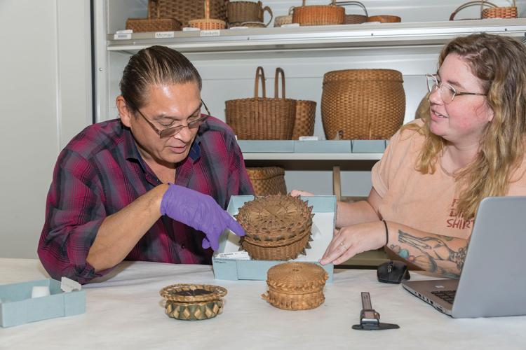 A man and a woman look at a basket. Behind them on shelves are many other baskets