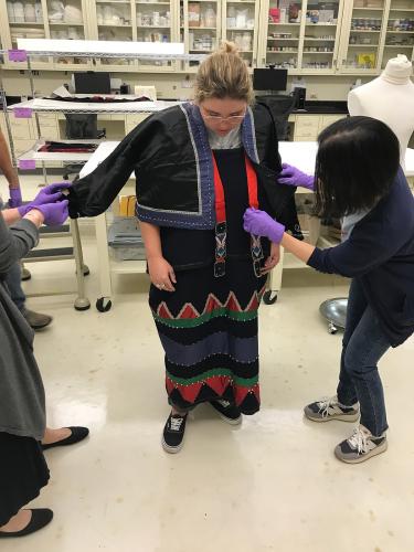 Two women help another get into a black and red wool skirt and jacket 