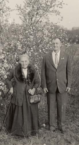 Isabel Meadows in a coat with a fur collar stands next to John Peabody Harrington wearing a suit in front of a tree
