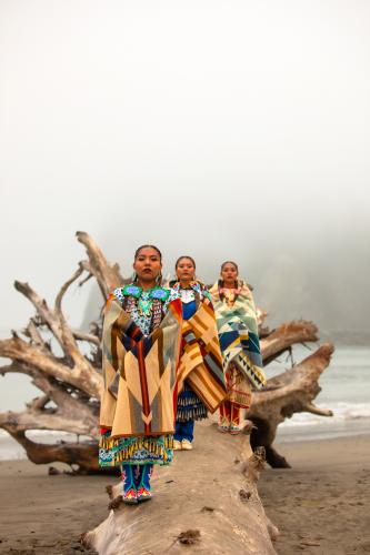 Three jingle dress dancers pose on a fallen tree log on the foggy shore.