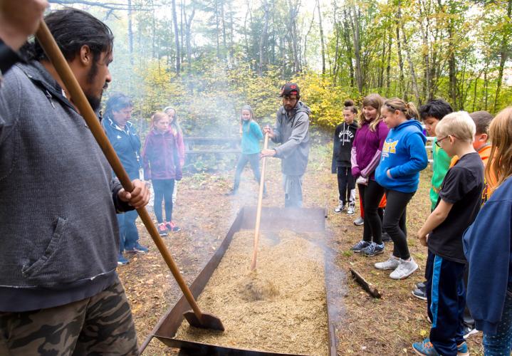 Two men stir wild rice while a group of kids watches.