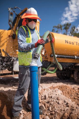 A worker in hard hat, reflective vest, and protective mask, filling a pipe with water from a hose connected to a tank on a truck.