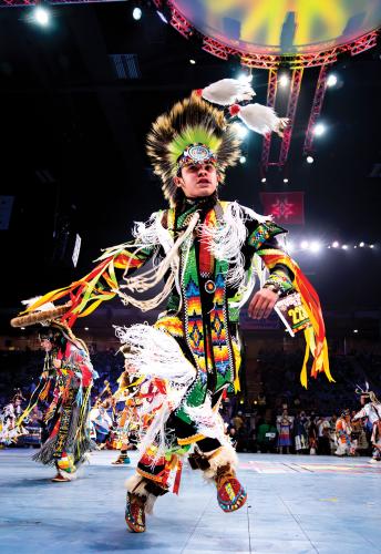 A male dancer wearing a colorful outfit with fringe, ribbons, and a headdress