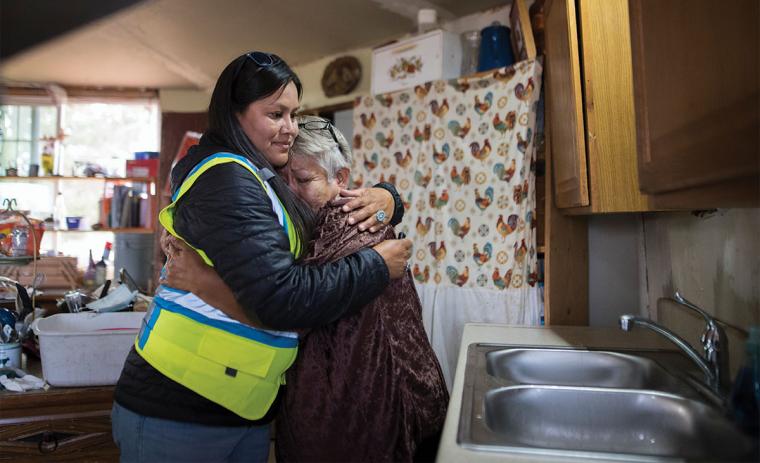 An older woman hugs a younger woman in a reflective vest near a kitchen sink