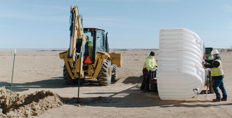 An excavator near a hold in the ground. Two workers stand nearby with a water cistern.