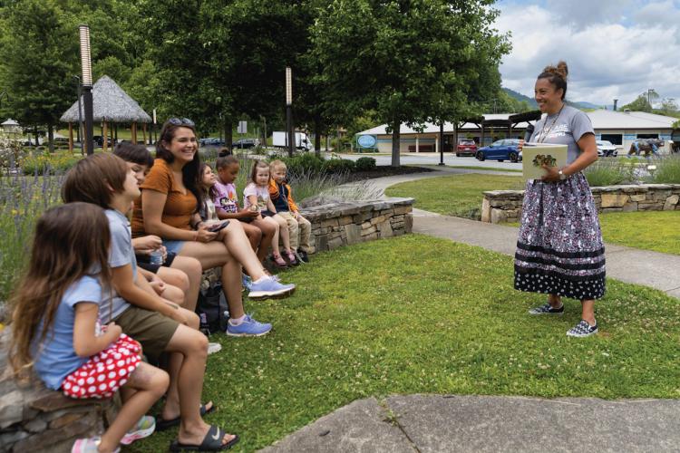 A smiling woman holds a book, facing a semi-circle of children seated on a rock wall outdoors
